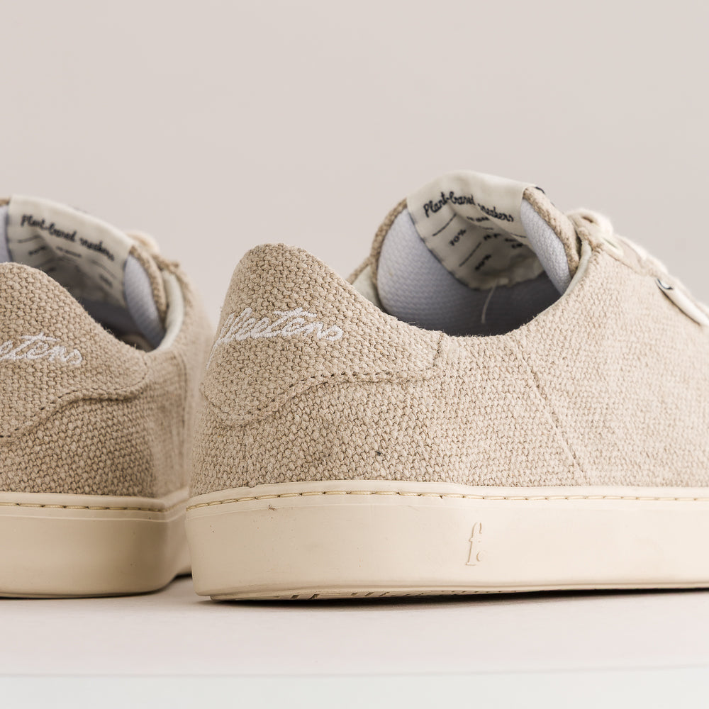 Close-up of a pair of light beige wool sneakers with white rubber soles and white laces, photographed against a plain light background.