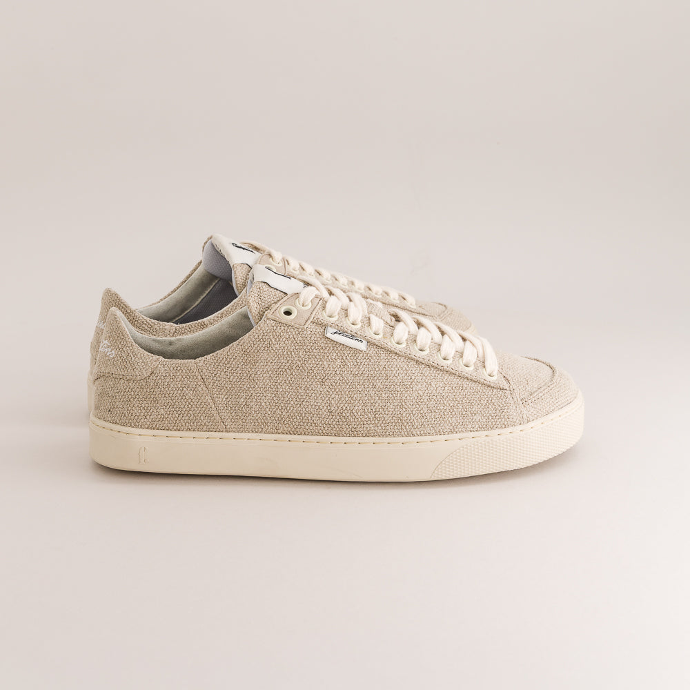 A pair of low-top wool barefoot sneakers in a light beige color, featuring white laces and white rubber soles, photographed against a neutral light gray background.