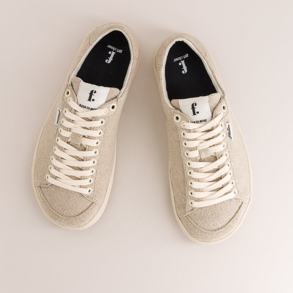 A pair of low-top wool barefoot sneakers in a light beige color, featuring cream-colored laces, white rubber soles, and a black interior with a white logo.
