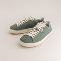 A pair of low-top wool sneakers in a muted sage green color, featuring off-white laces and thick off-white rubber soles, photographed on a light beige studio background.