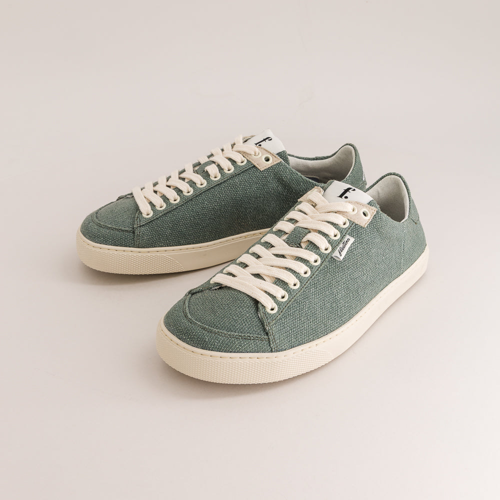 A pair of low-top wool sneakers in a muted sage green color, featuring off-white laces and thick off-white rubber soles, photographed on a light beige studio background.
