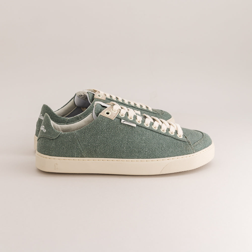 A pair of low-top canvas sneakers in a muted sage green color, featuring off-white laces and a thick off-white rubber sole, photographed against a neutral light gray background.