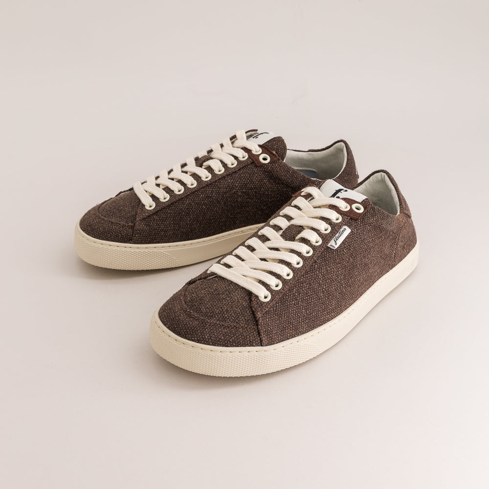 A pair of low-top wool sneakers in dark brown with off-white laces and soles, displayed on a light beige studio background.