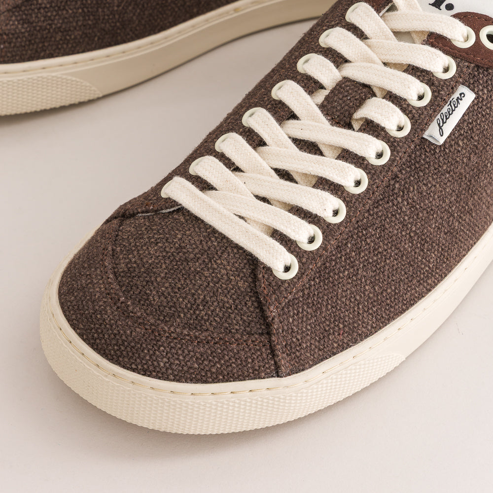 A close-up of a brown wool low-top sneaker with white laces and a cream-colored rubber sole, featuring a small white 'Fiction' brand tag on the side.