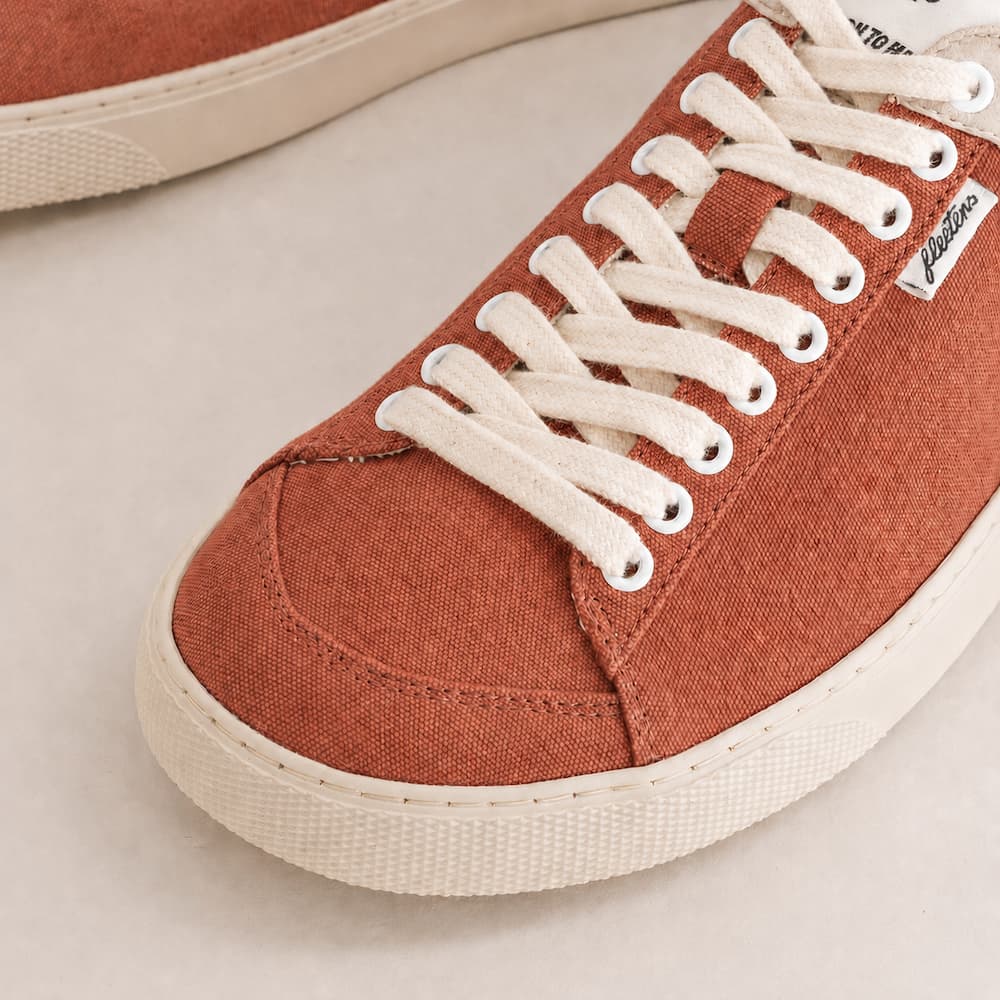 Close-up of a low-top canvas sneaker in a warm rust orange color with white laces and a white rubber sole, on a light beige background.