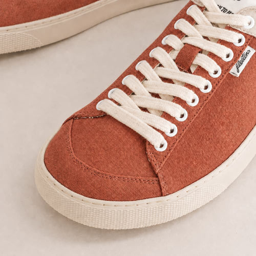 Close-up of a low-top canvas sneaker in a warm rust orange color with white laces and a white rubber sole, on a light beige background.