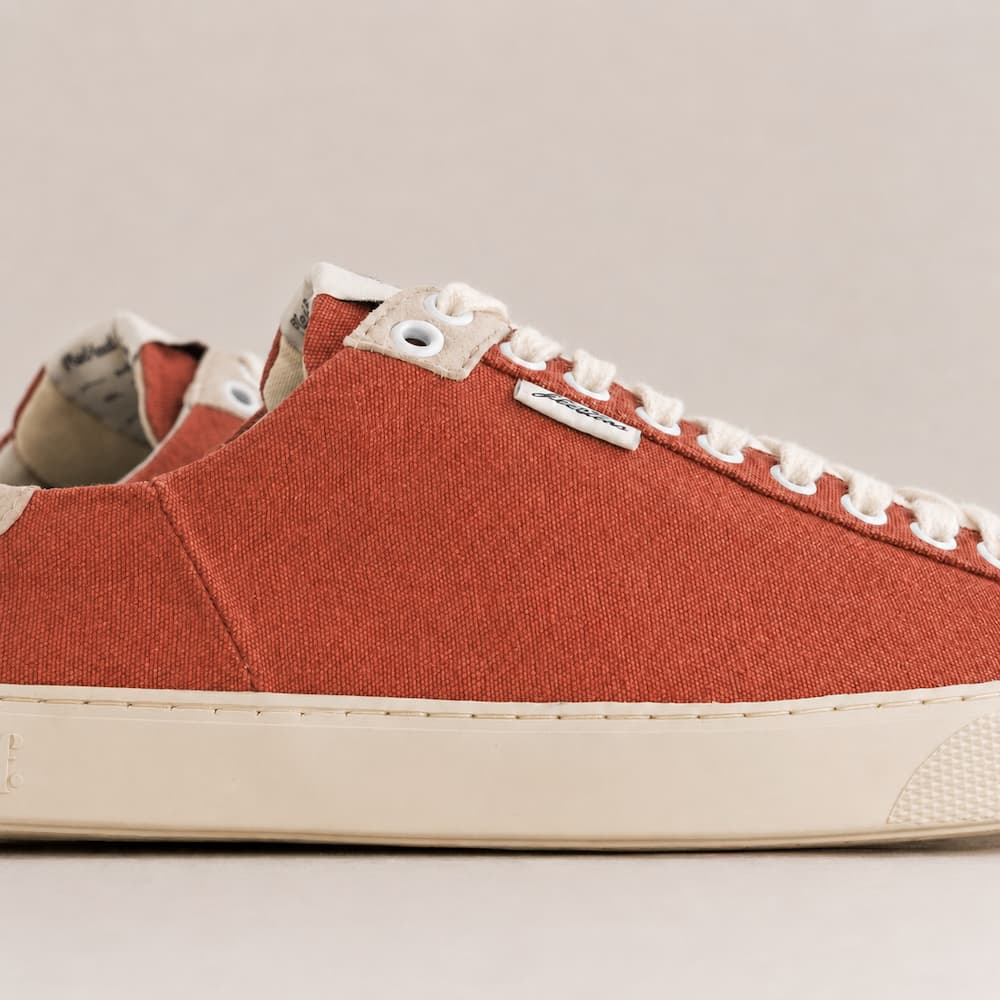 A close-up side view of a low-top canvas sneaker in terracotta red with white laces and a cream-colored rubber sole, shown against a neutral beige background.