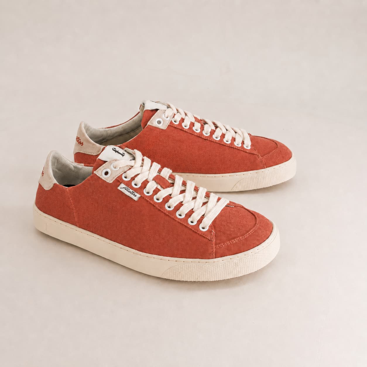 A pair of low-top canvas sneakers in a warm rust orange color, featuring white laces and white rubber soles, photographed on a light beige studio background.