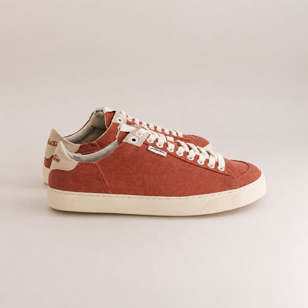 A pair of low-top canvas sneakers in a terracotta rust color with off-white laces and a cream-colored sole, shown from a side profile on a light gray background.