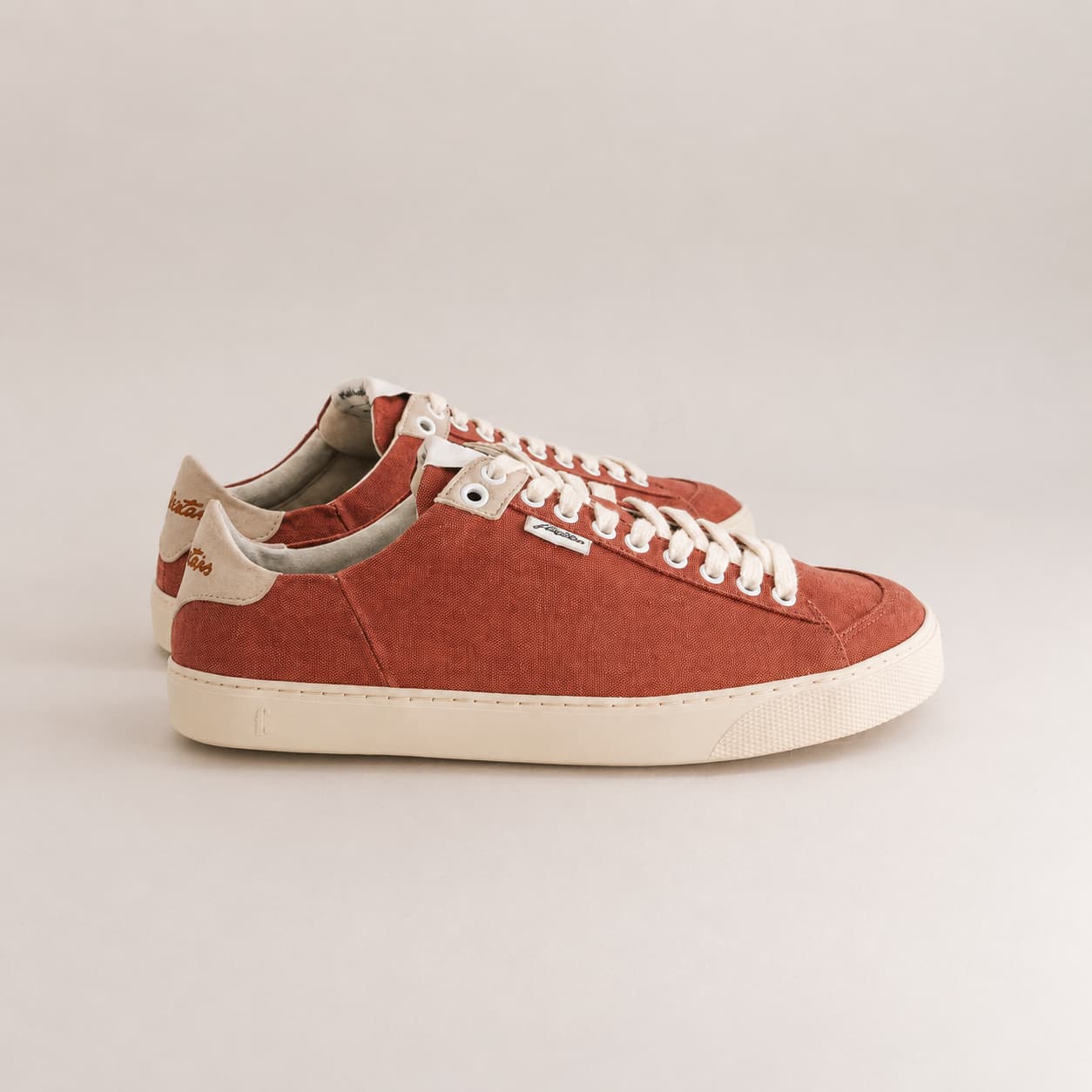 A pair of low-top canvas sneakers in a terracotta rust color with off-white laces and a cream-colored sole, shown from a side profile on a light gray background.