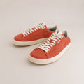 A pair of low-top canvas sneakers in a warm rust-orange color, featuring off-white laces and thick off-white rubber soles, displayed on a light beige background.