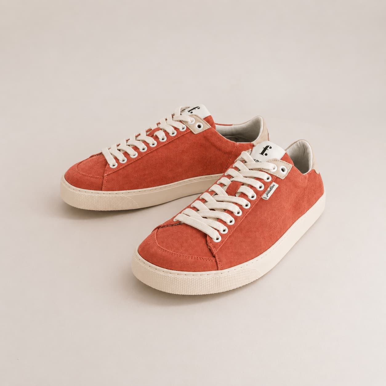 A pair of low-top canvas sneakers in a warm rust-orange color, featuring off-white laces and thick off-white rubber soles, displayed on a light beige background.