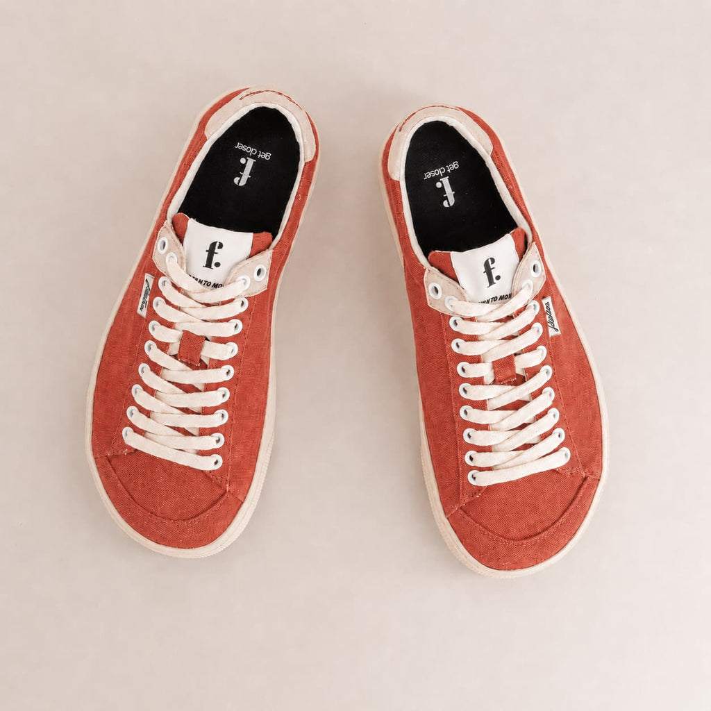 A pair of low-top canvas barefoot sneakers in a muted terracotta red color, featuring white laces, white rubber soles, and a black interior with a white logo.