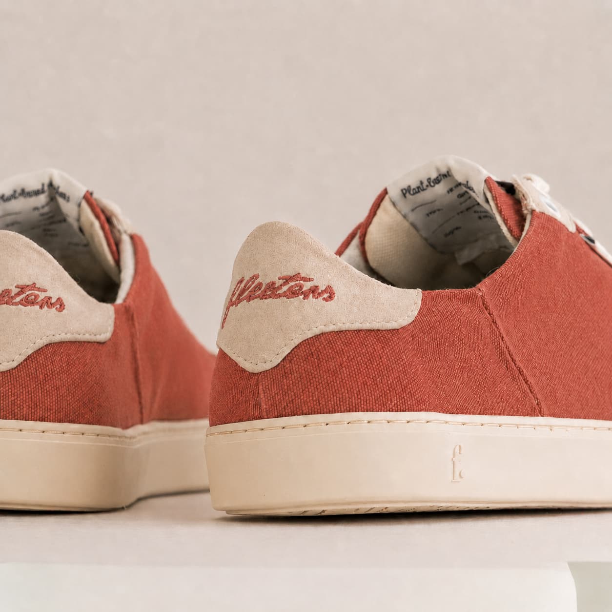 Close-up of a pair of low-top sneakers showing the heel and side panels. The shoes have a muted terracotta red canvas upper with a contrasting warm beige heel tab and white rubber sole.