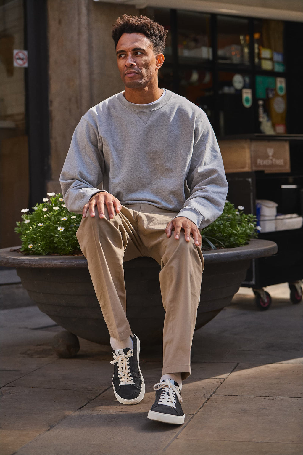 A man sitting on a large outdoor planter wearing a light gray crewneck sweatshirt, beige trousers, and black barefoot sneakers with white laces and soles, set against a blurred urban storefront background.