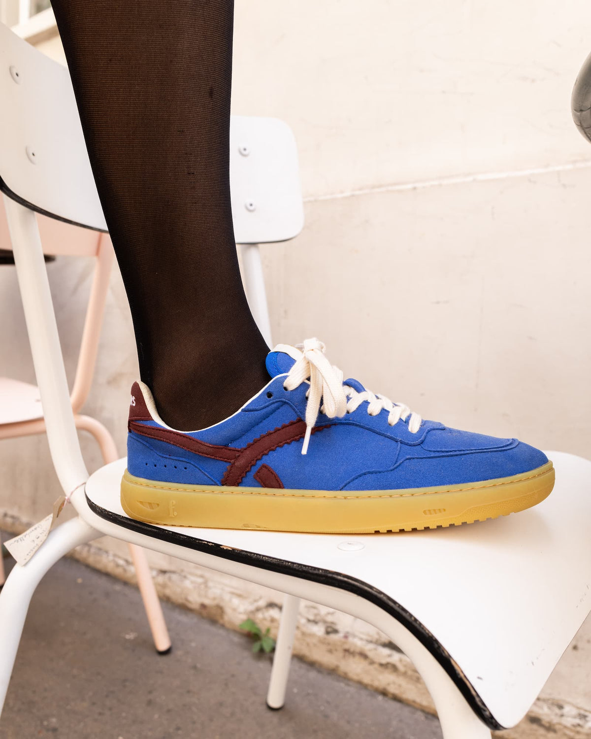 A blue suede barefoot sneaker with a brown side stripe, white laces, and a yellow gum sole, shown on a foot wearing black tights while sitting on a white chair.