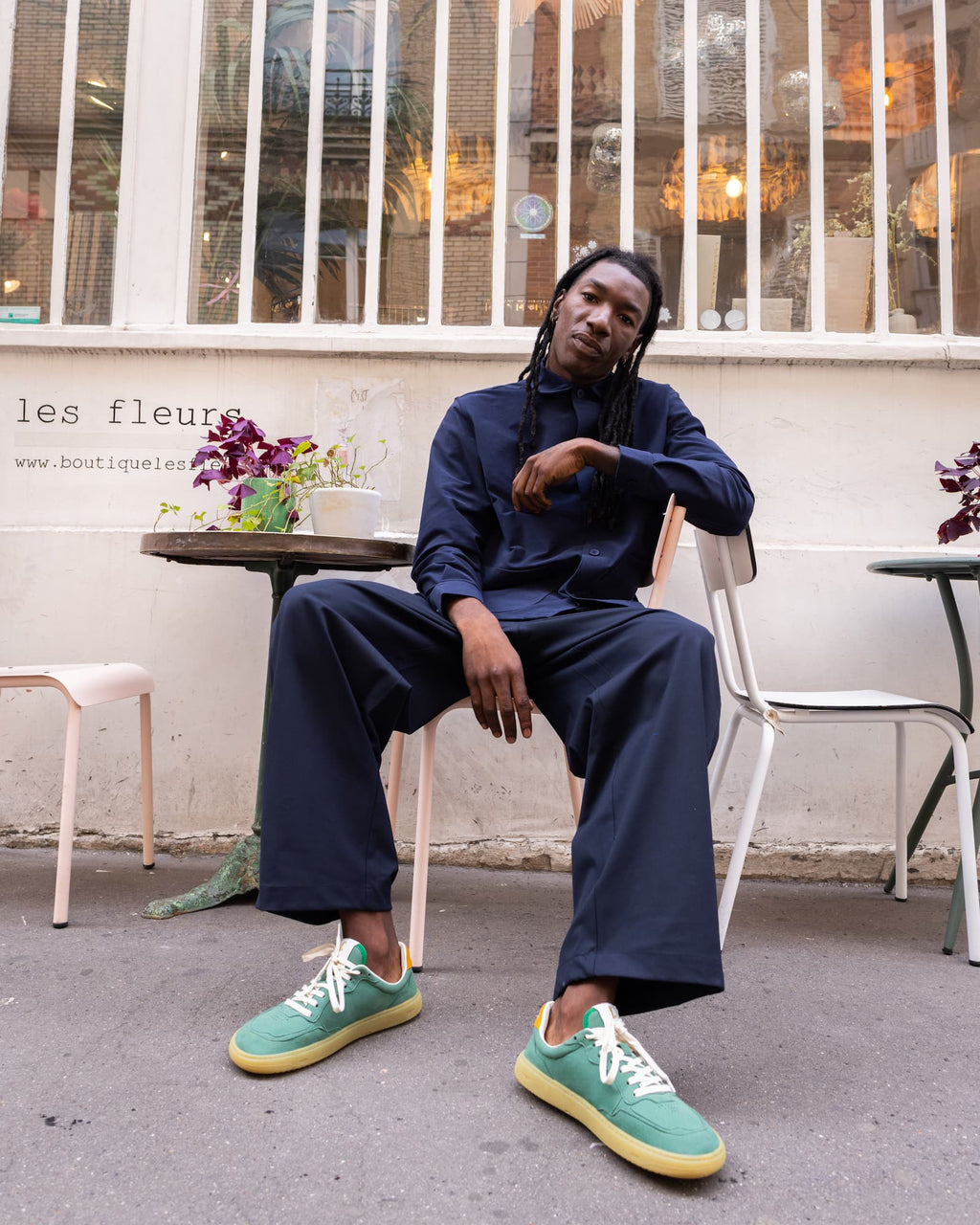 A man sits on a white chair in an outdoor cafe setting, wearing a matching navy blue long-sleeve top and wide-leg trousers with mint green and yellow barefoot sneakers.