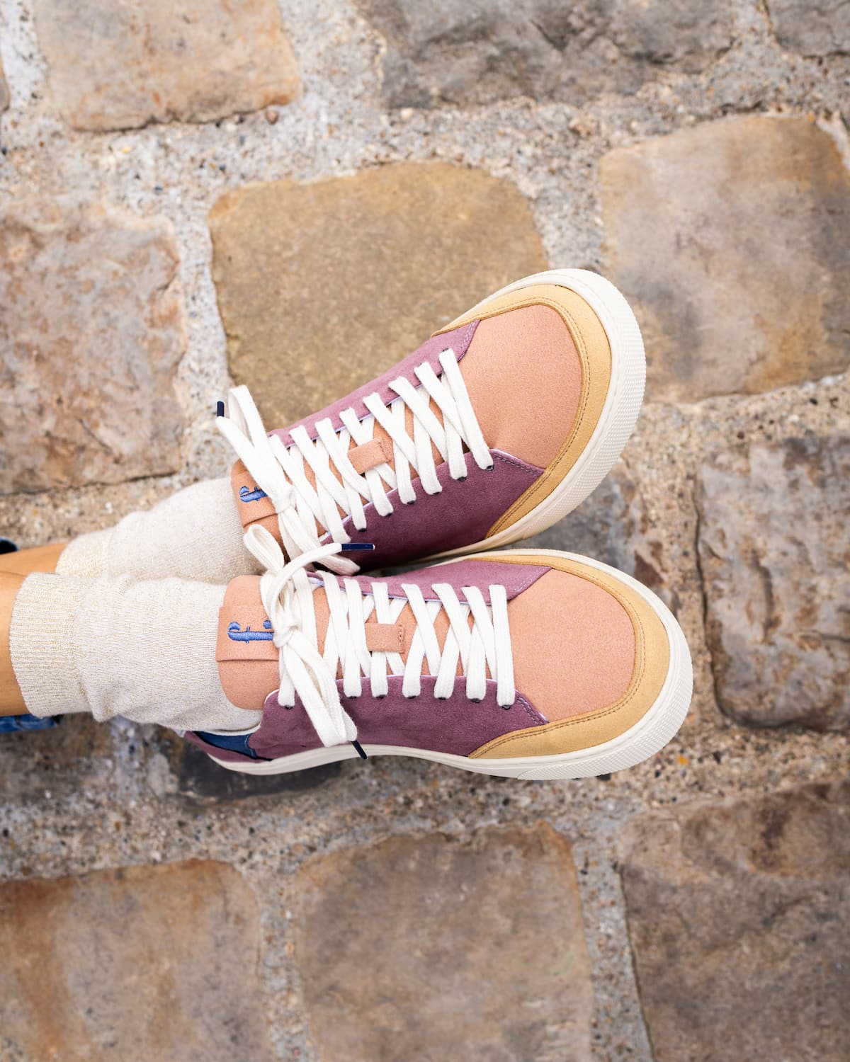 A pair of low-top barefoot sneakers with a mauve-purple and peach canvas upper, white laces, and cream soles, worn with white socks on a cobblestone surface.