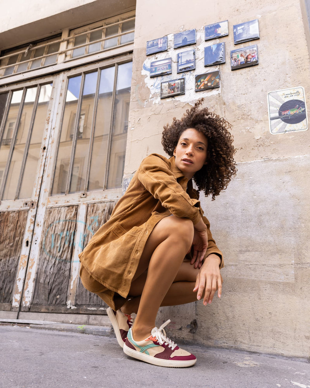 A woman with curly hair crouches against a concrete wall in an urban setting, wearing a brown corduroy jacket and multicolor barefoot sneakers with burgundy, teal, and white panels.