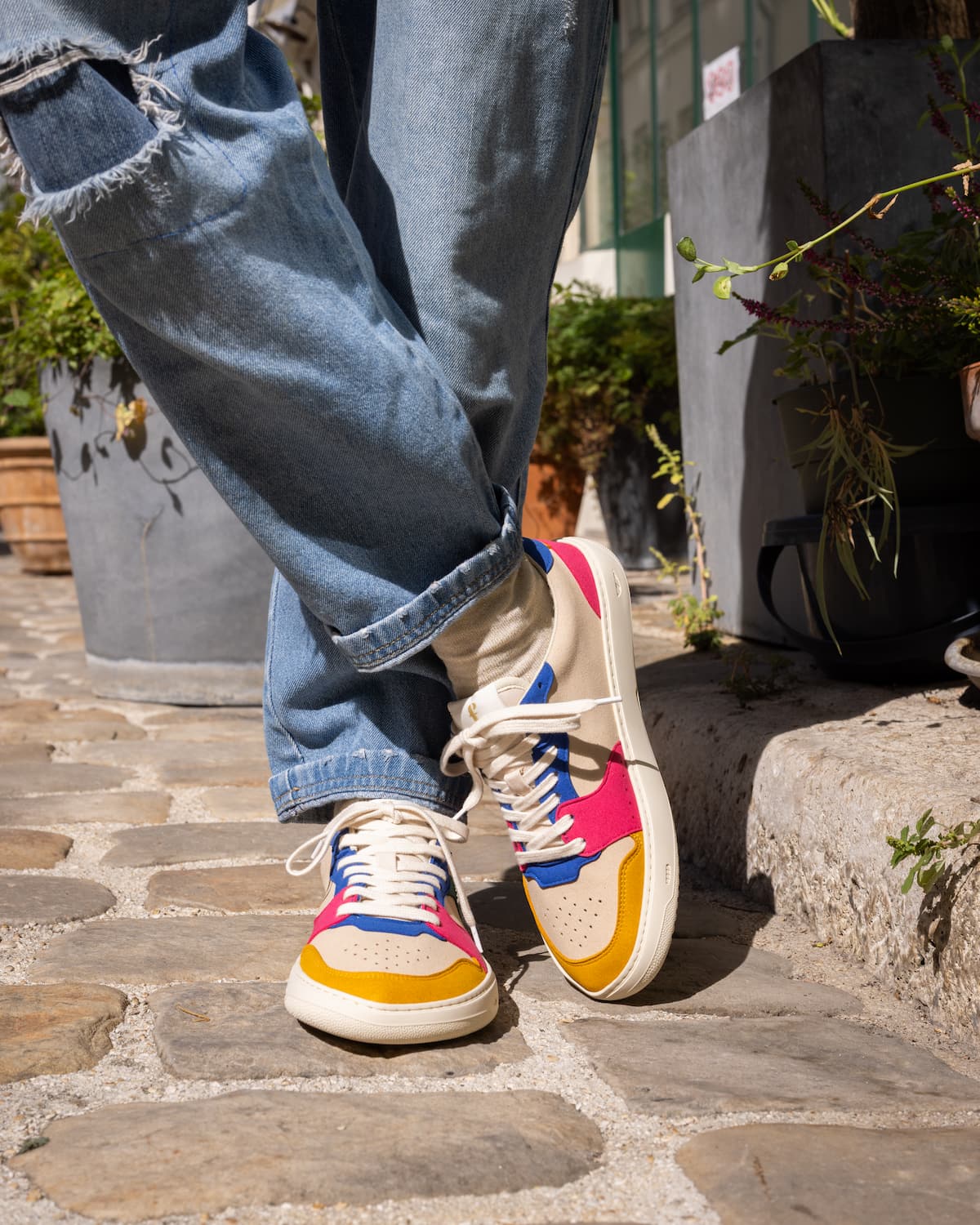 A person wearing colorful low-top barefoot sneakers with ripped blue jeans, standing on a stone patio outdoors.