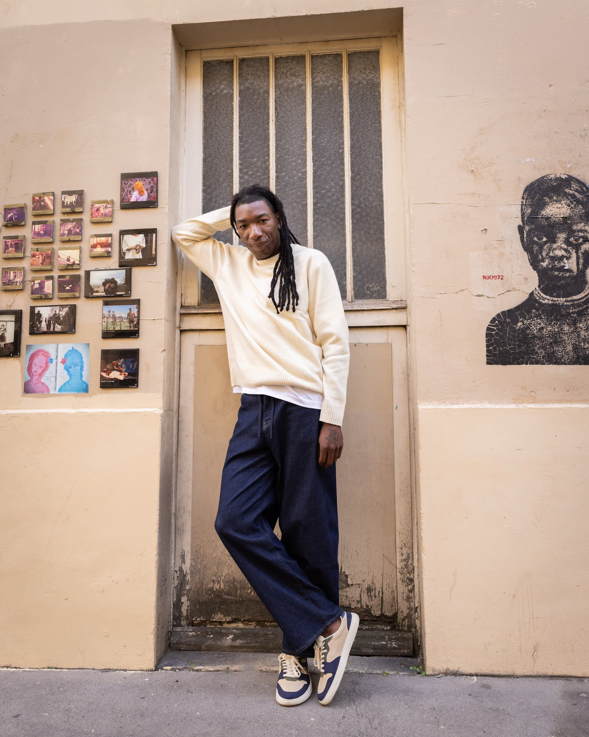 A man with dreadlocks stands leaning against a beige wall in front of a barred door, wearing a cream long-sleeve shirt, navy wide-leg pants, and white barefoot sneakers with navy accents. The wall is decorated with small framed photos and a black portrait print.