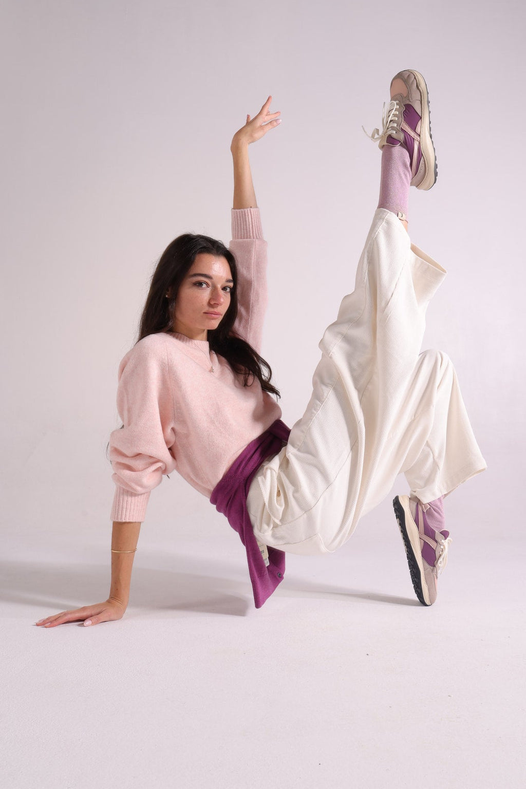 A woman in a studio setting wearing a light pink sweatshirt, cream wide-leg pants, and purple socks, posed dynamically on a white floor with one leg raised high.