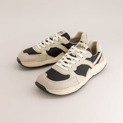 A pair of retro-style low-top barefoot sneakers in beige, black, and off-white, featuring white laces and a chunky cream midsole, photographed on a light beige studio background.
