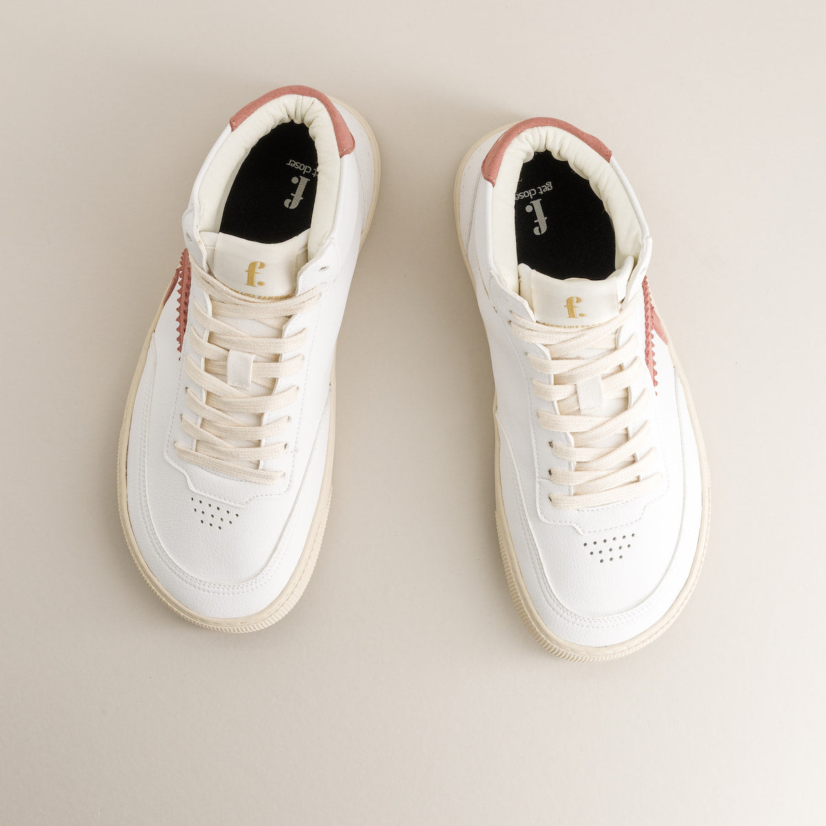 Pair of white mid-ankle barefoot sneakers with pink accents on a beige background