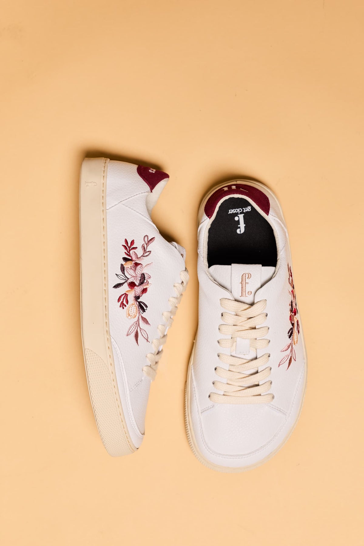 A pair of white low-top barefoot sneakers with floral embroidery on the sides, shown from top and side angles on a solid beige background. The shoes feature cream laces, a black interior lining, and a burgundy heel tab.