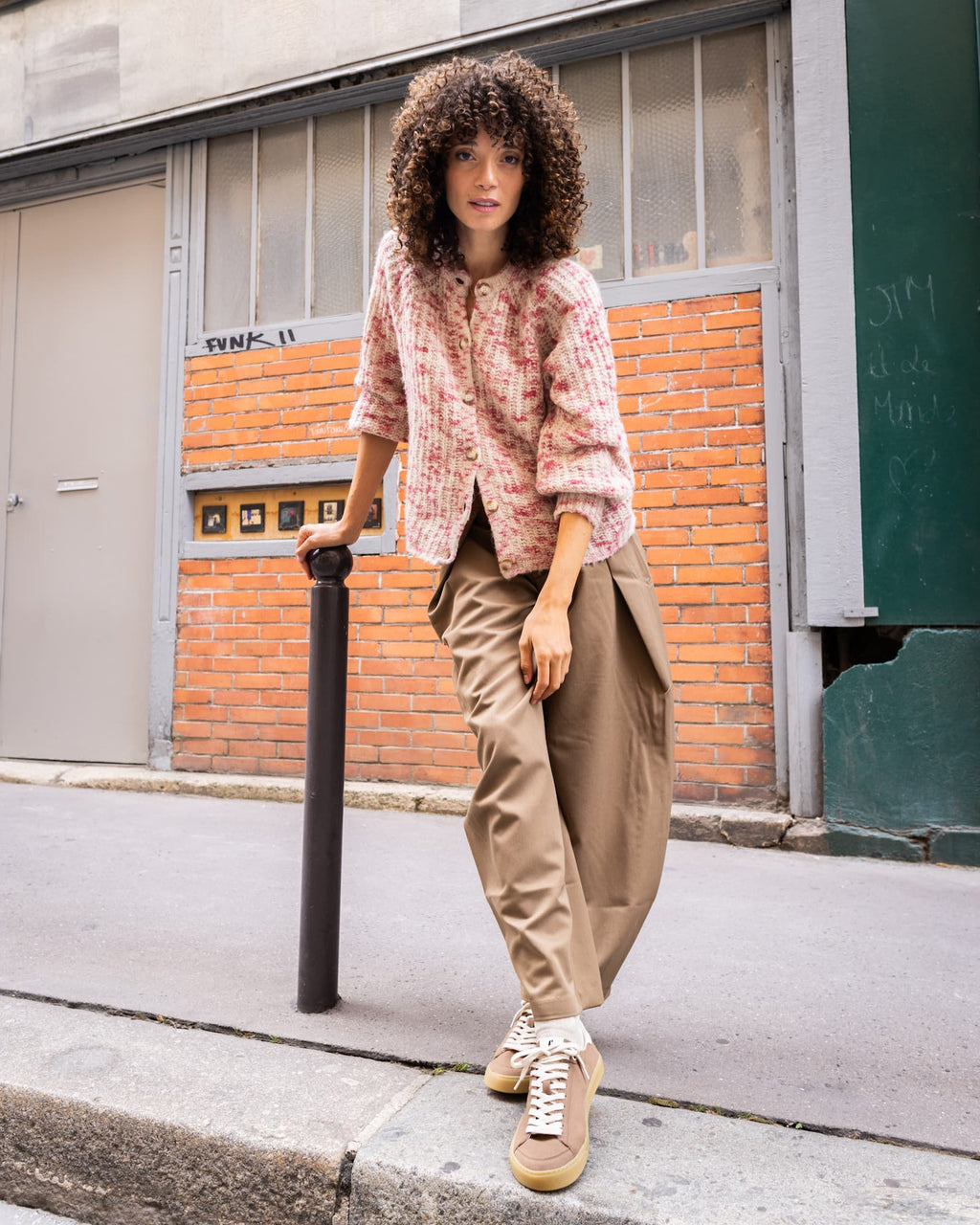 A woman stands on a city sidewalk in front of a brick building, wearing a pink and white textured jacket, beige wide-leg trousers, and beige barefoot sneakers with white laces.