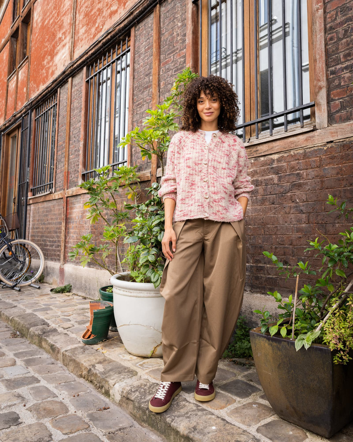 A woman stands outdoors on a cobblestone street in front of a brick building, wearing a light pink floral long-sleeve blouse, brown wide-leg trousers, and burgundy minimalist sneakers with gold soles.