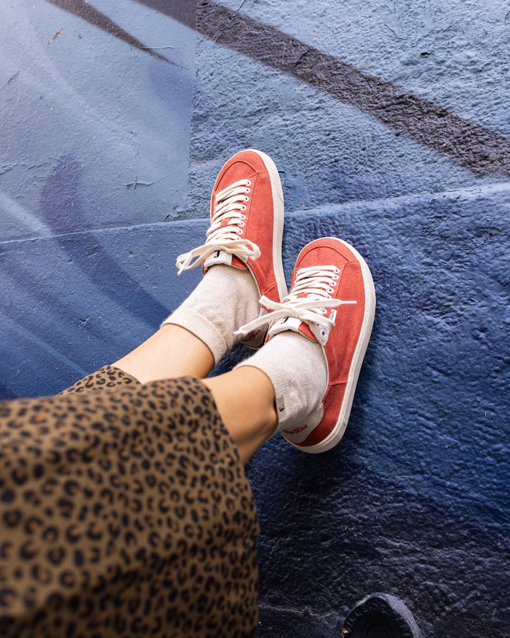 A person wearing red low-top barefoot sneakers with white laces and white socks, standing on a dark blue textured wall. A leopard-print garment is visible at the lower left.