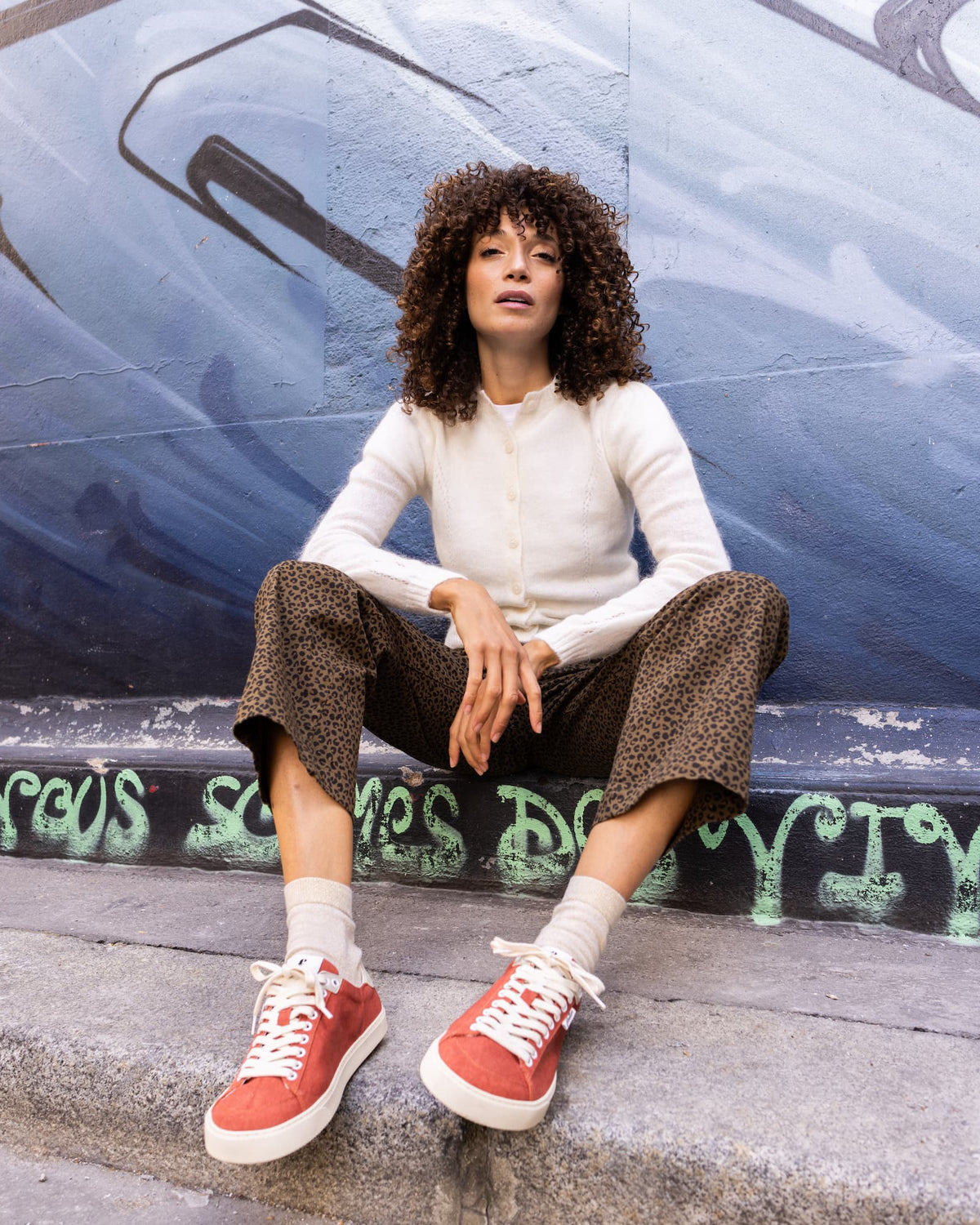 A woman with curly hair sits on concrete steps in front of a graffiti-covered wall, wearing a cream ribbed cardigan, brown patterned cropped pants, white socks, and red low-top barefoot sneakers.