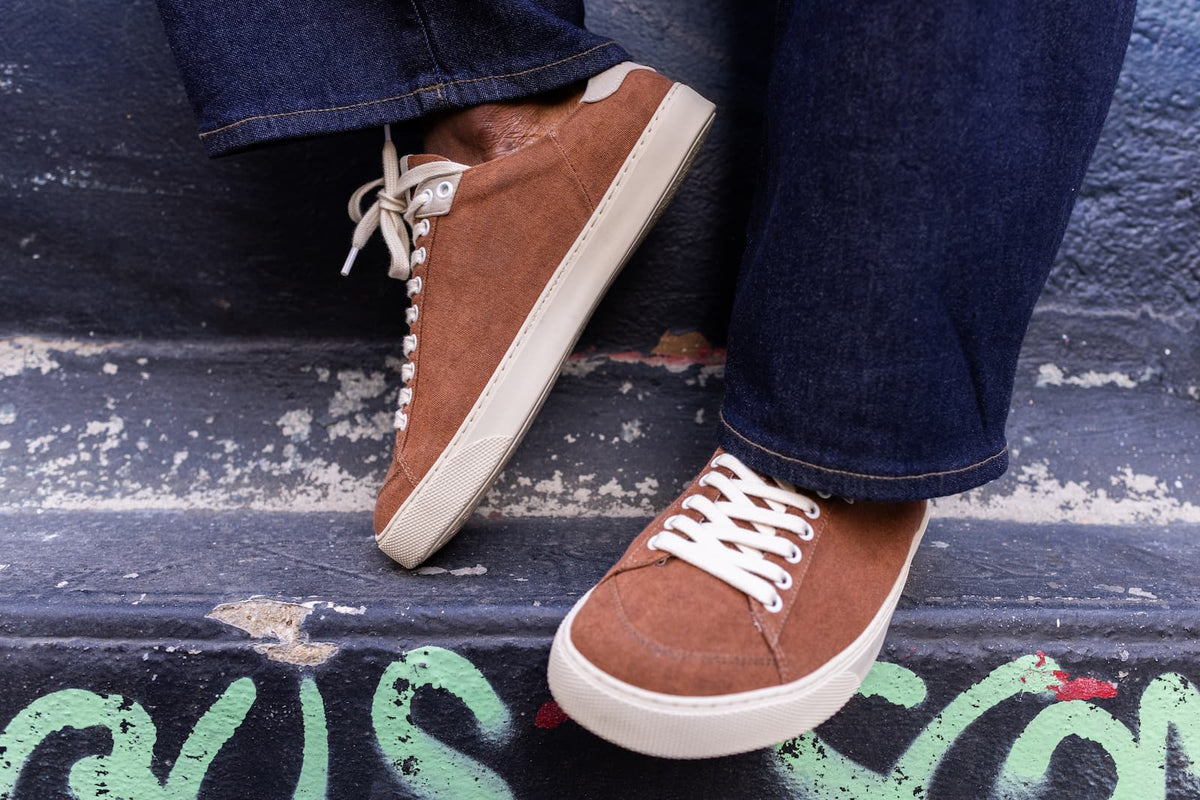 A close-up of a person wearing brown suede barefoot sneakers with white laces and white soles, standing on dark painted steps with graffiti.