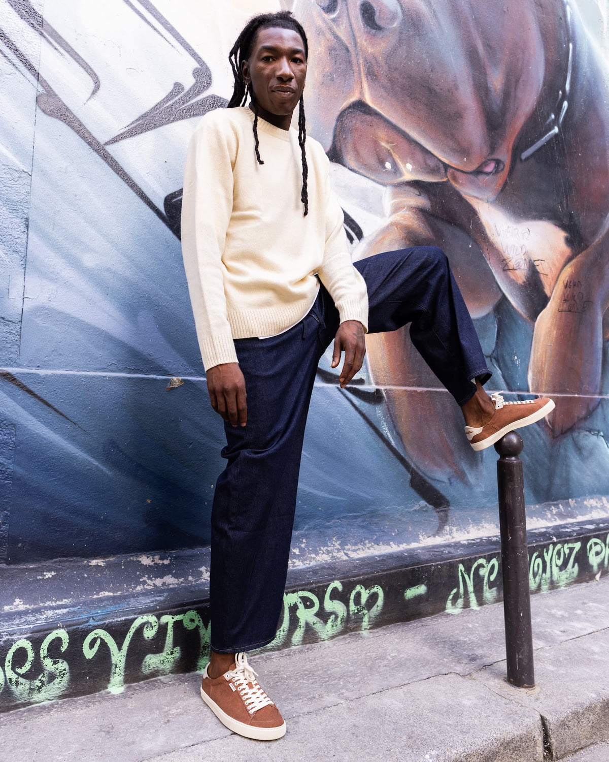 A man wearing a cream knit sweater, dark navy trousers, and brown barefoot sneakers stands against a large graffiti mural.