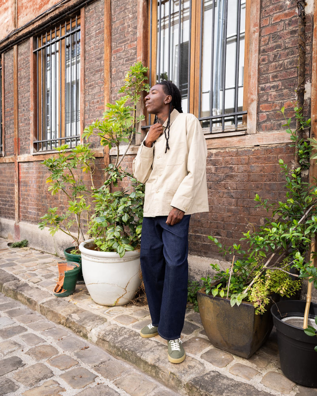 A man wearing a cream-colored long-sleeve shirt, navy blue trousers, and green barefoot sneakers stands on a cobblestone street in front of a brick building with potted plants.