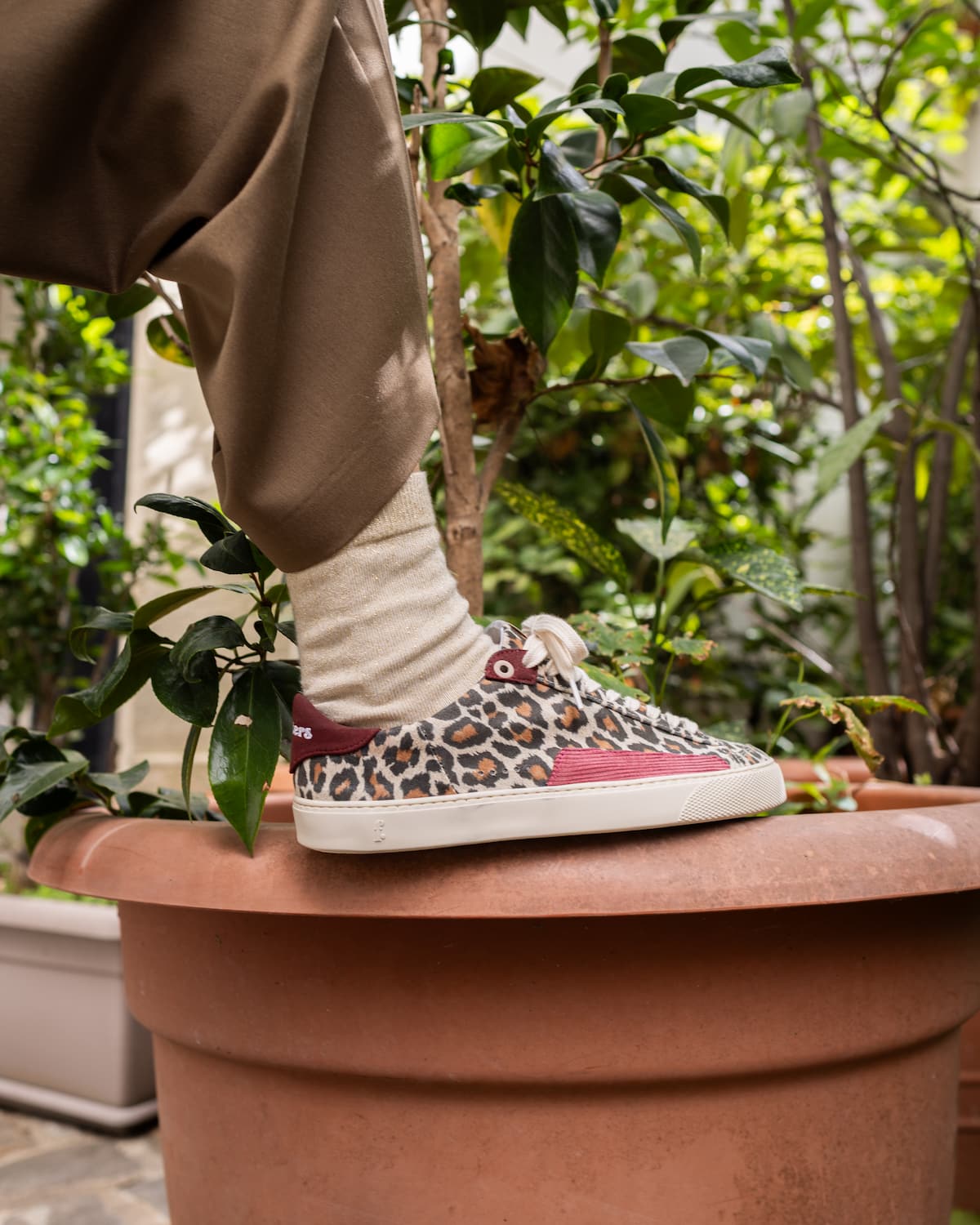 A person wearing brown pants and a cream sock stands beside a large terracotta planter, showcasing a low-top barefoot sneaker with a leopard print pattern, a pink side stripe, and a white rubber sole in an outdoor garden setting.