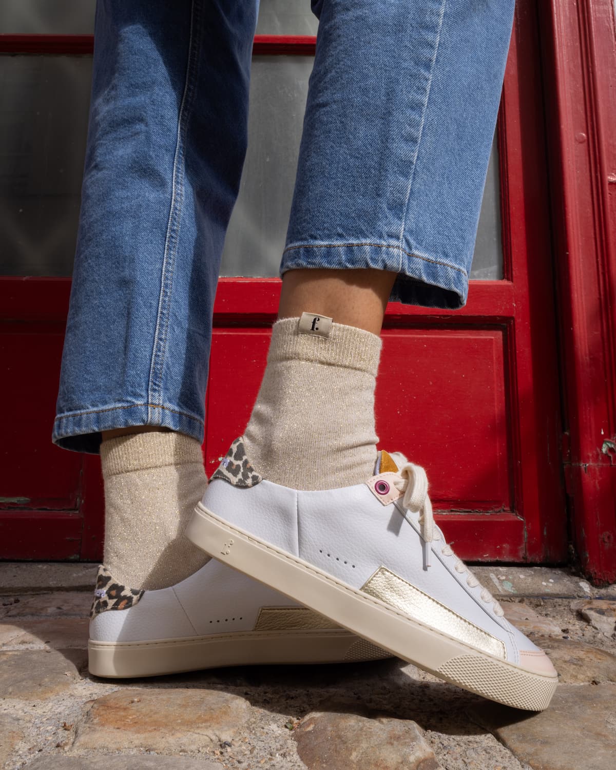 A person wearing blue jeans, beige crew socks, and white low-top barefoot sneakers with leopard-print detailing near the heel, standing outdoors in front of a red Parisian door.