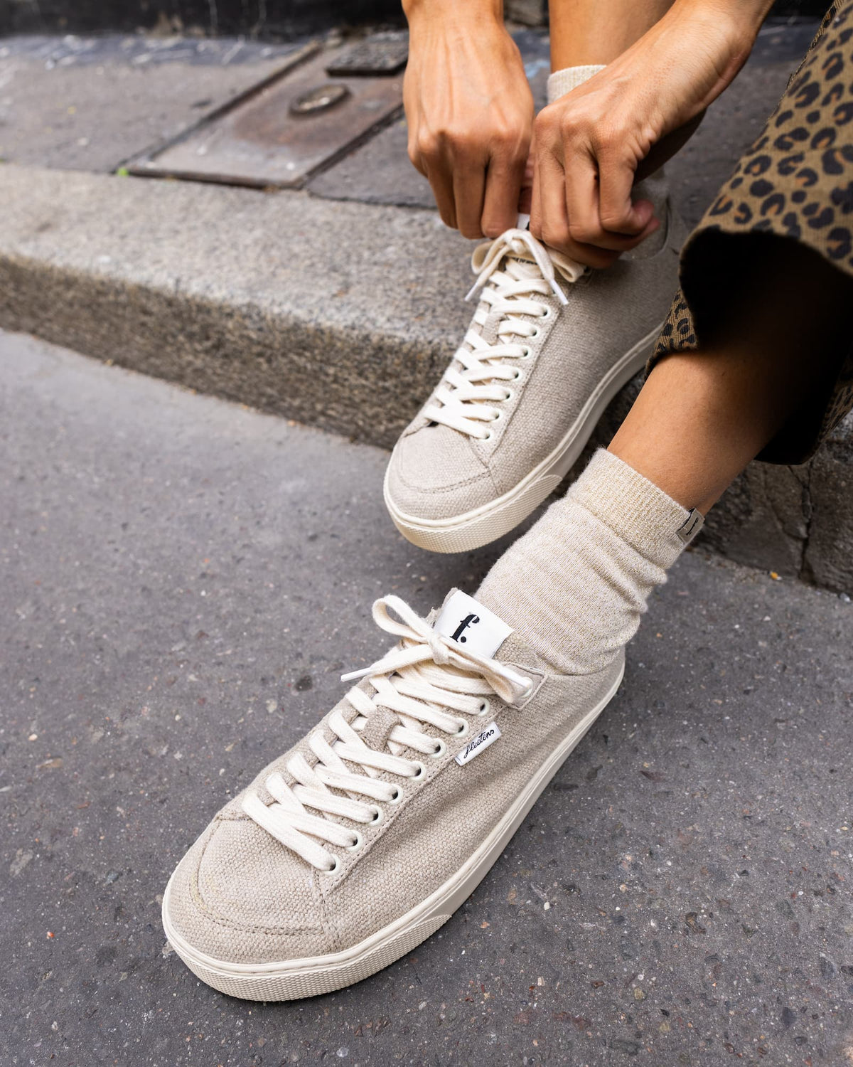 A person sitting outdoors on a curb tying the laces of a pair of grey wool barefoot sneakers with beige socks. The shoes have white rubber soles and a small white brand tag on the side.
