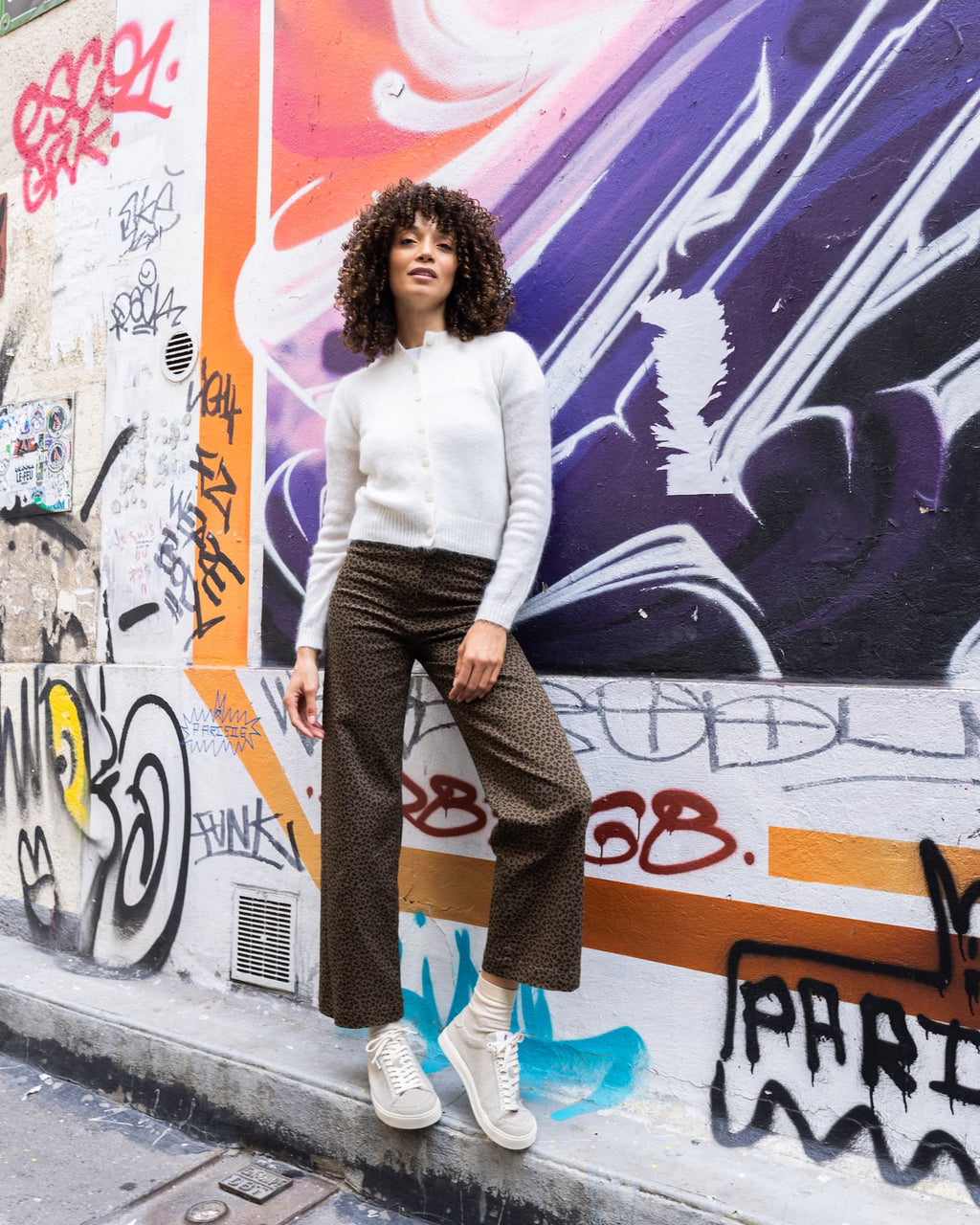 A woman stands against a graffiti-covered wall wearing a white ribbed knit cardigan, brown patterned wide-leg cropped pants, and grey wool barefoot sneakers.