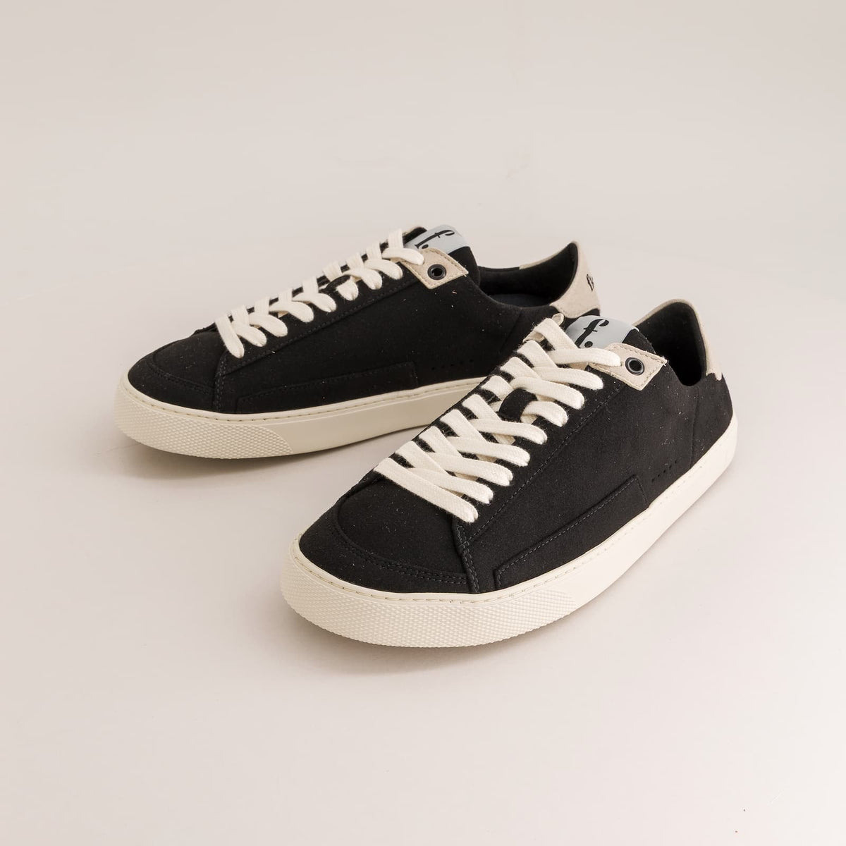A pair of black canvas low-top barefoot sneakers with off-white laces and off-white rubber soles, displayed on a clean light gray studio background.