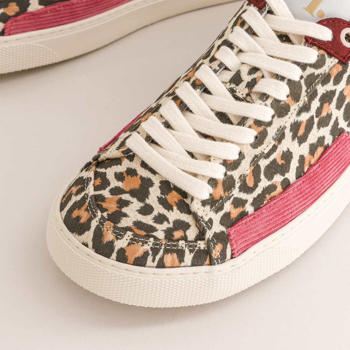 Close-up of a low-top barefoot  sneaker with a leopard print pattern, white laces, and a pink side stripe, shown on a light neutral background.