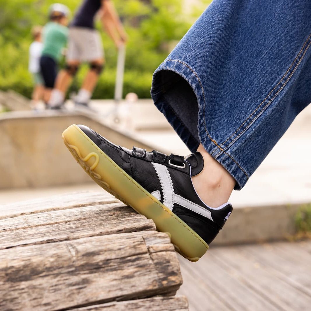A close-up of a person's foot wearing a black skate barefoot shoe with white side stripes and a gum sole, stepping onto a wooden ledge at an outdoor skatepark.
