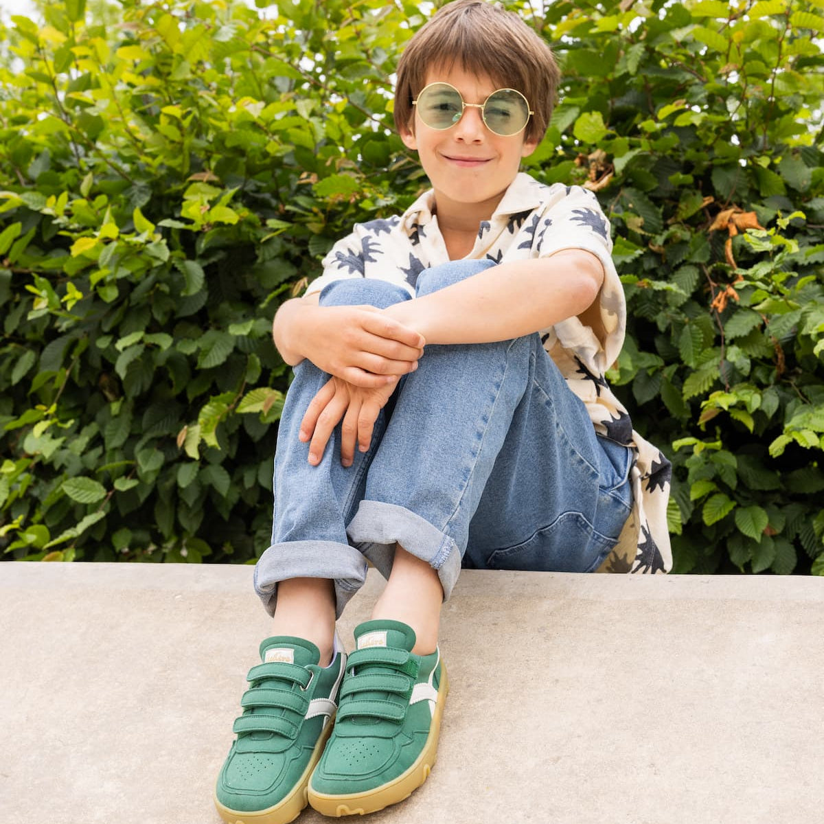 A young boy wearing round green-tinted sunglasses, a white patterned short-sleeve shirt, blue jeans, and green sneakers sits on a concrete ledge with a green hedge background.
