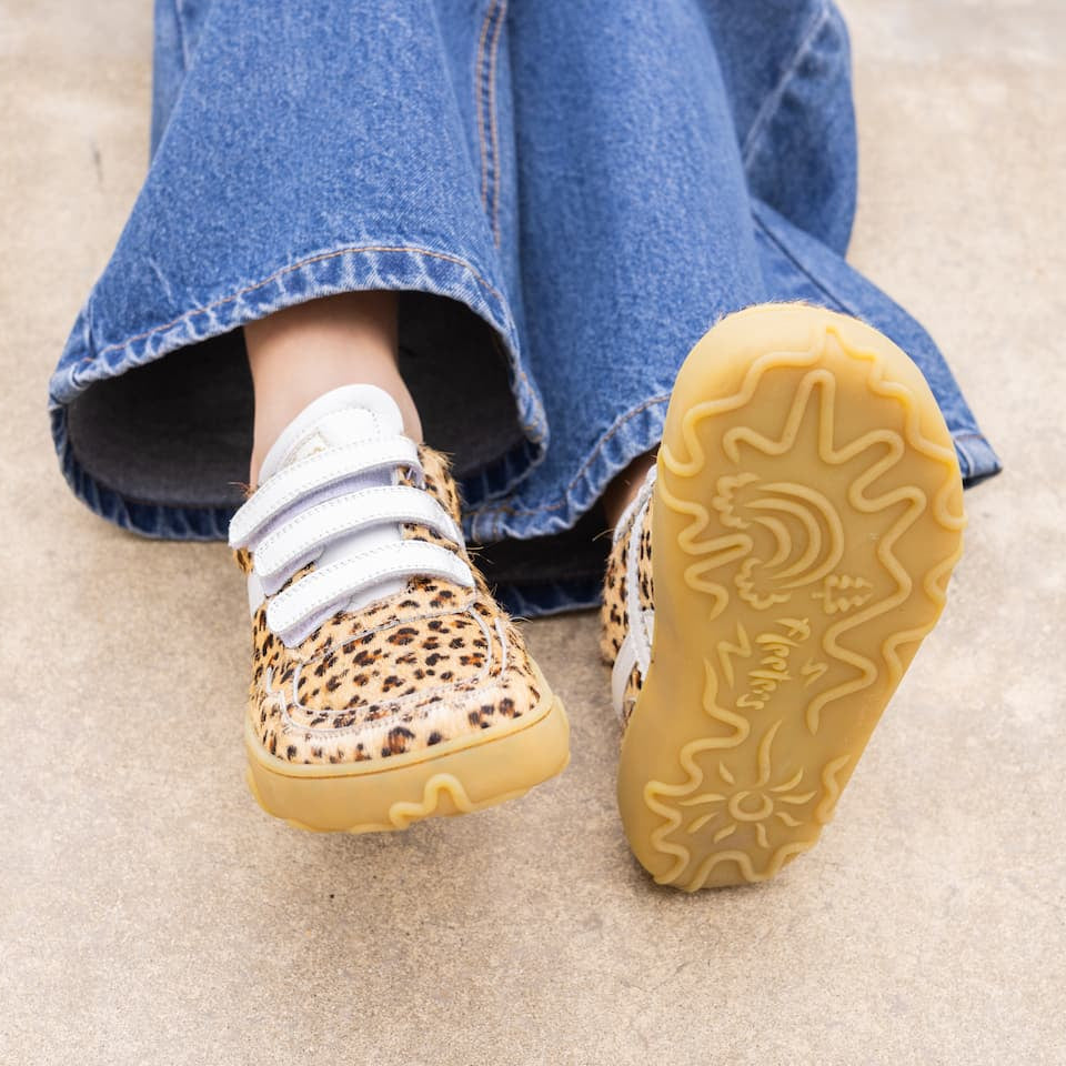 A close-up of a person's legs wearing blue jeans and leopard-print slip-on shoes with white elastic laces, shown against a light concrete floor. The sole of one shoe is raised to show the tread pattern.
