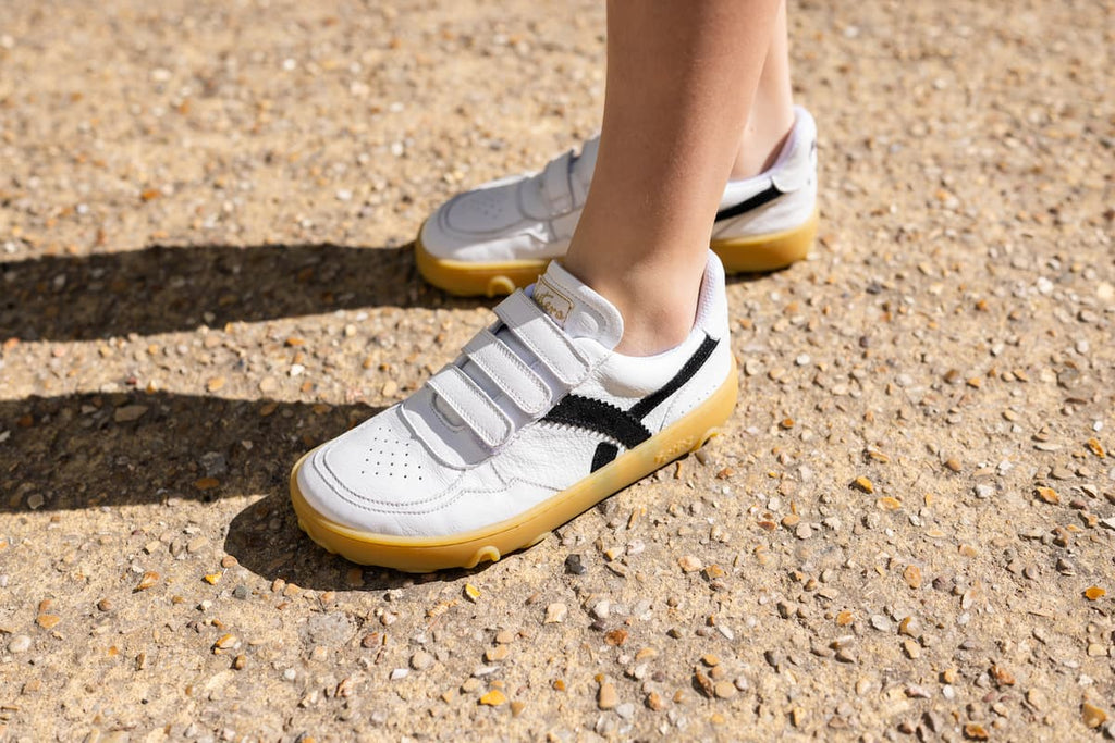 White barefoot sneakers with hook-and-loop straps, black side accents, and gum soles, shown being worn outdoors on a gravel surface.
