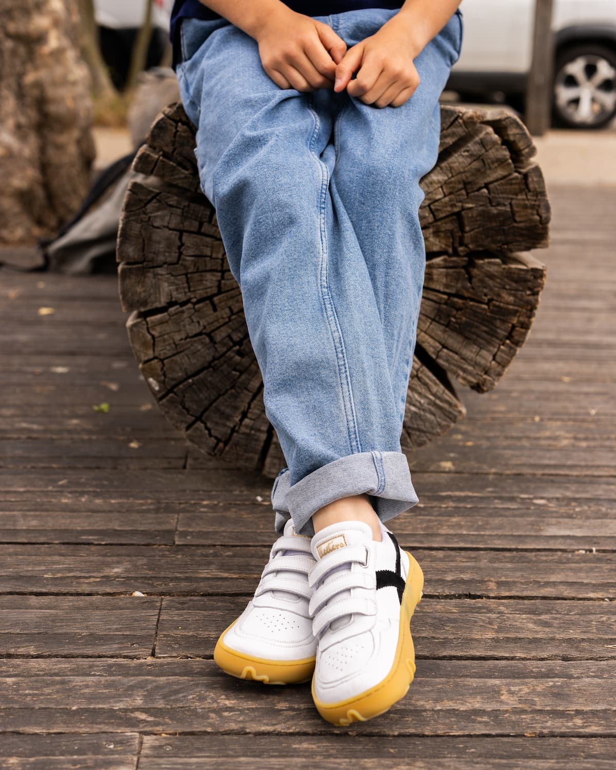 A child wearing light blue jeans and white barefoot sneakers with yellow soles sits on a tree stump on a wooden deck.