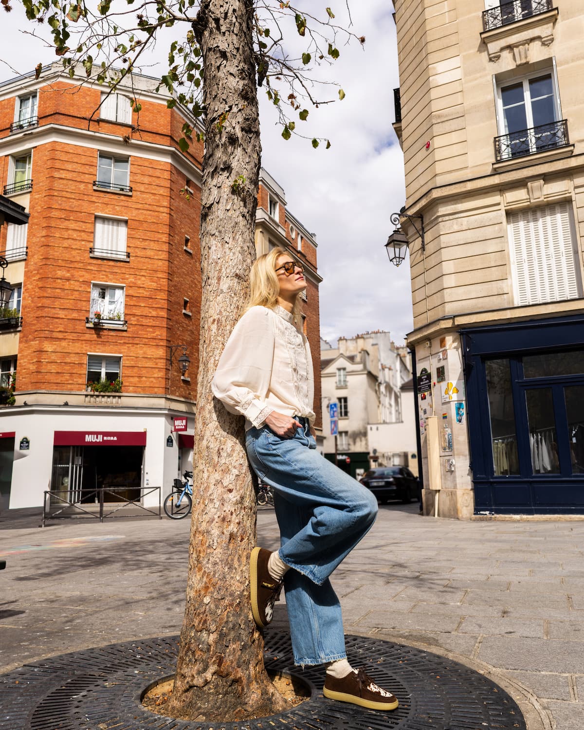 A woman wearing a cream blouse, blue jeans, and brown barefoot shoes leans against a tree in an outdoor urban street setting.
