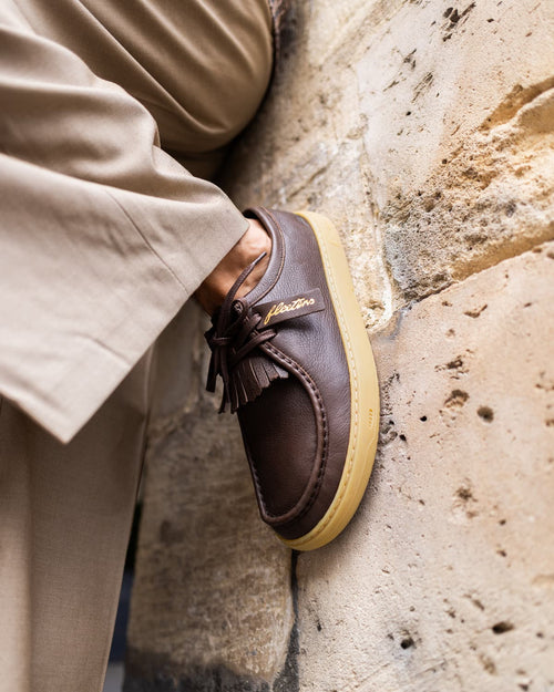 A close-up of a person wearing a brown leather tassel barefoot loafer with a yellow sole, leaning against a textured stone wall.