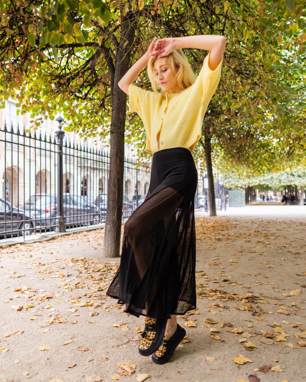 A woman posing outdoors on a tree-lined street, wearing a pale yellow short-sleeve button-up shirt, a high-waisted black midi skirt with sheer side panels, and barefoot loafers with leopard print detailing.
